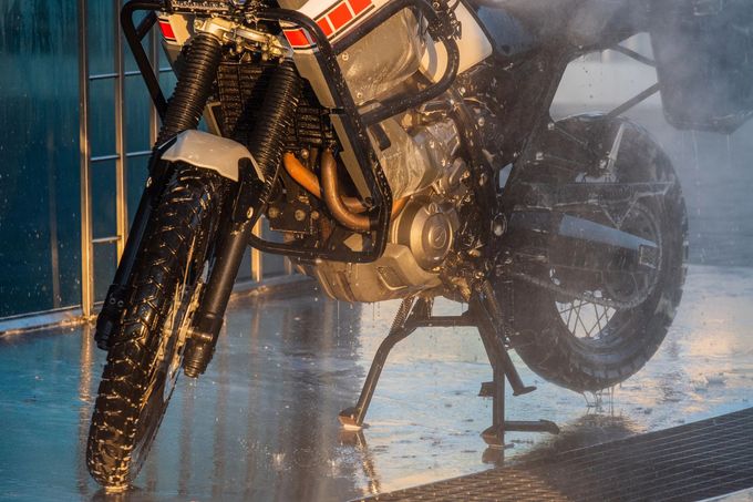 Motocicletta parcheggiata sotto la pioggia, ruota anteriore bagnata e motore visibile, con acqua che schizza tutt'intorno.