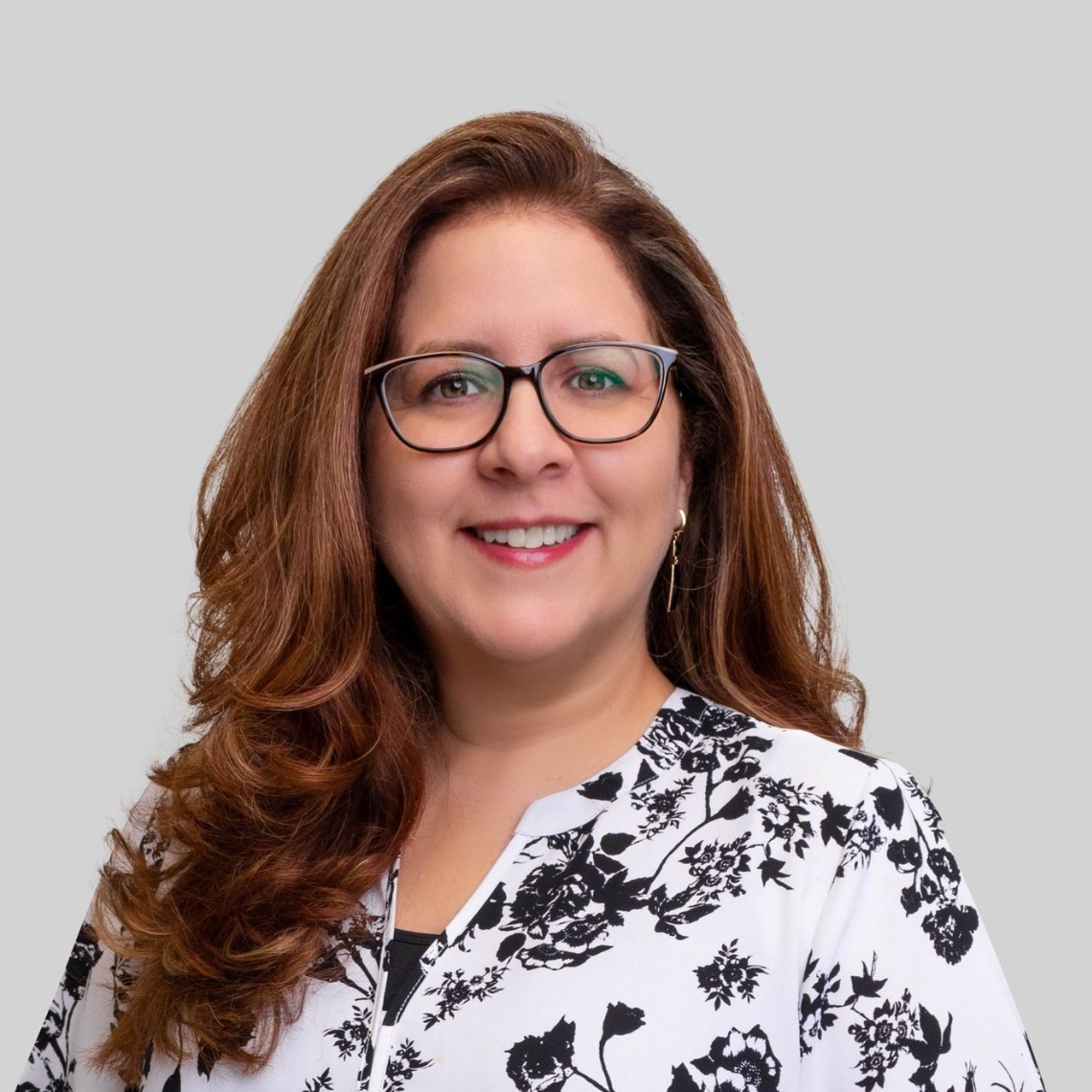 Woman with glasses, smiling, wearing a black and white floral print top, against a grey background. Dion Brady
