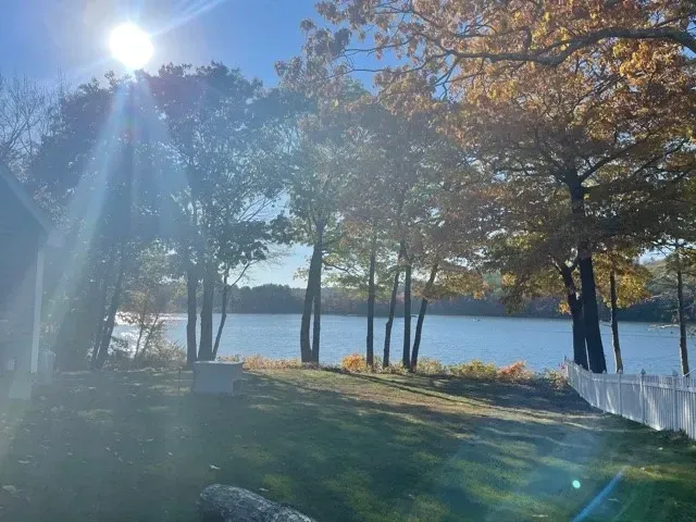 Sun shining over a lake, trees with yellow leaves, and a white fence.