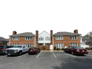 Apartment building with parked cars in front and white trim, viewed from a parking lot.