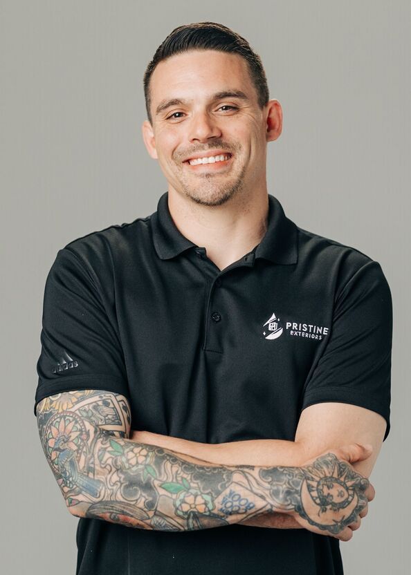Smiling person in a black polo with tattooed arms crossed against a gray background