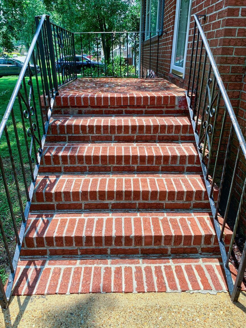 Brick front steps with black metal railings beside a red brick house and trees.
