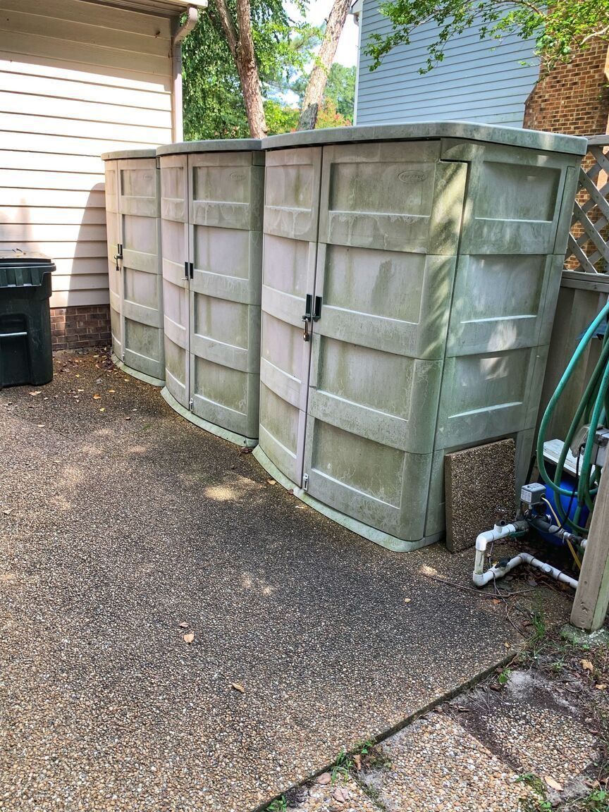 Large gray storage shed beside a house on a gravel driveway, with a black bin and blue bike nearby