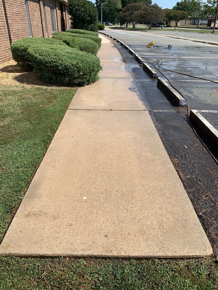 Sidewalk beside a brick building, with trimmed bushes on the left and a parking lot on the right.