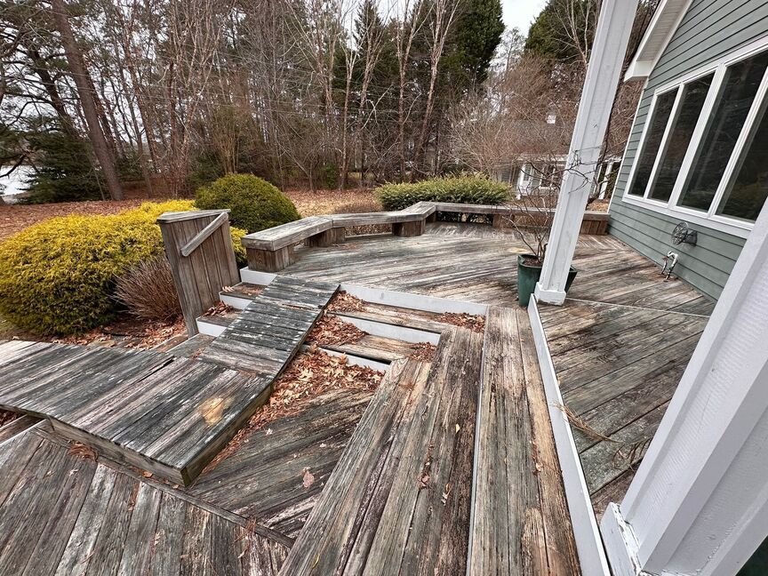 Damaged weathered wooden deck beside a house, with broken boards and overgrown shrubs nearby