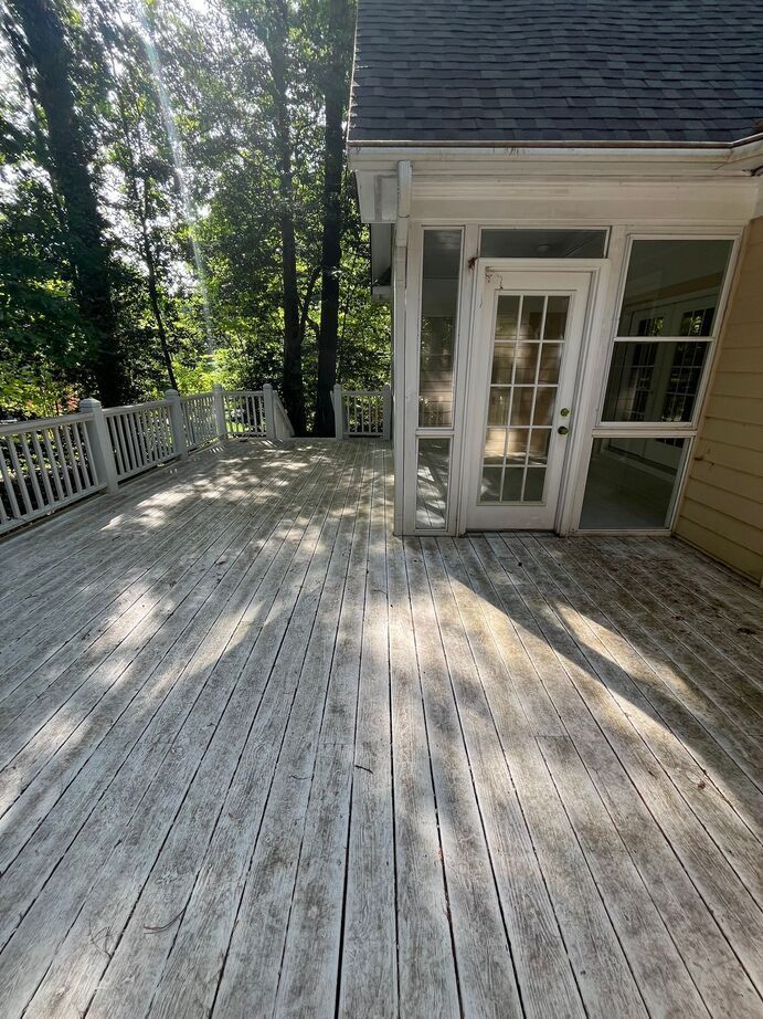 White wooden deck with railings beside a house and screened door, lit by sunlight and surrounded by trees