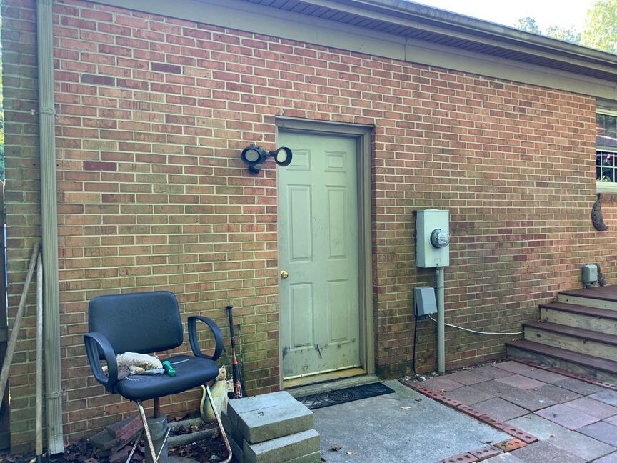Brick wall with a green door, wall light, utility meter, chair, and outdoor steps on a patio.