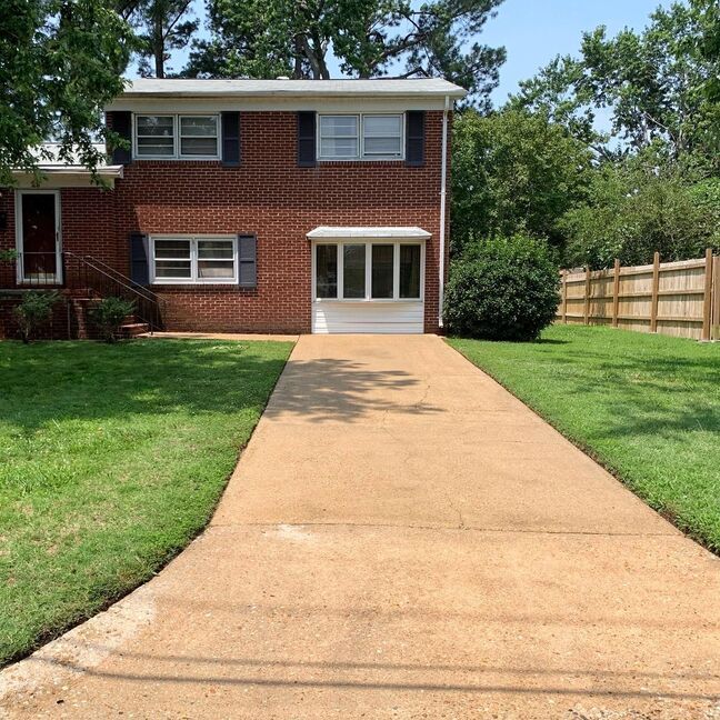 Brick two-story house with a concrete driveway, green lawn, and wooden fence under trees