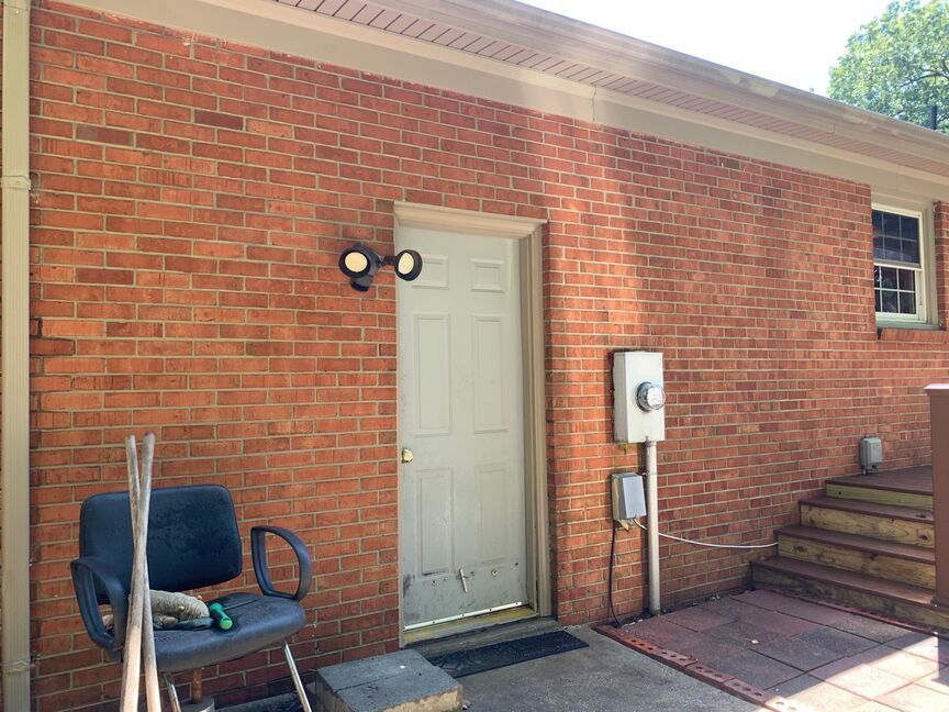 Brick patio with a cream door, wall light, utility meter, chair, and steps beside a red brick house