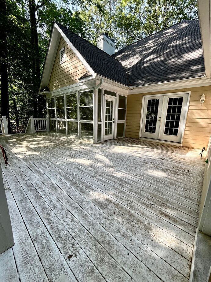Wooden porch and patio beside a light-colored house with large windows, set among trees in bright sunlight