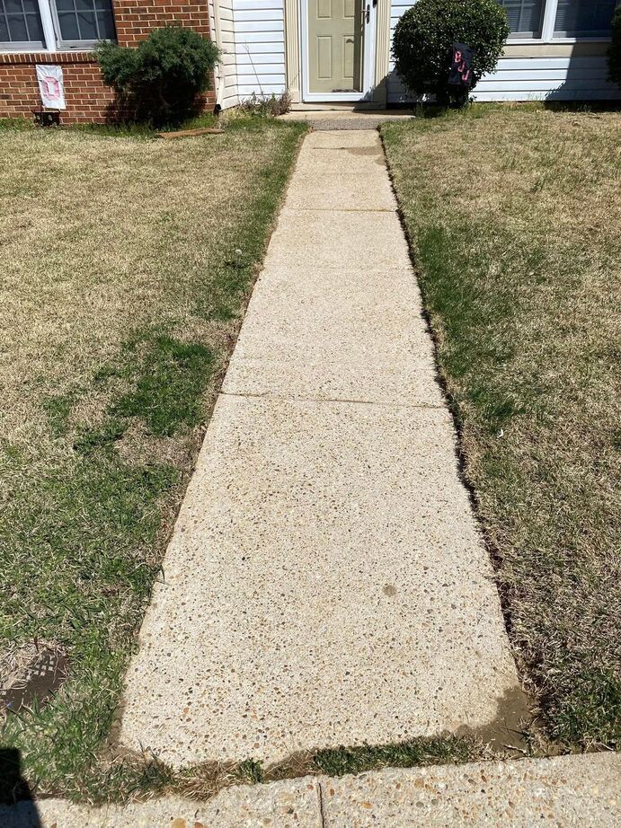 Concrete walkway leading to a house entrance through a patchy front lawn.