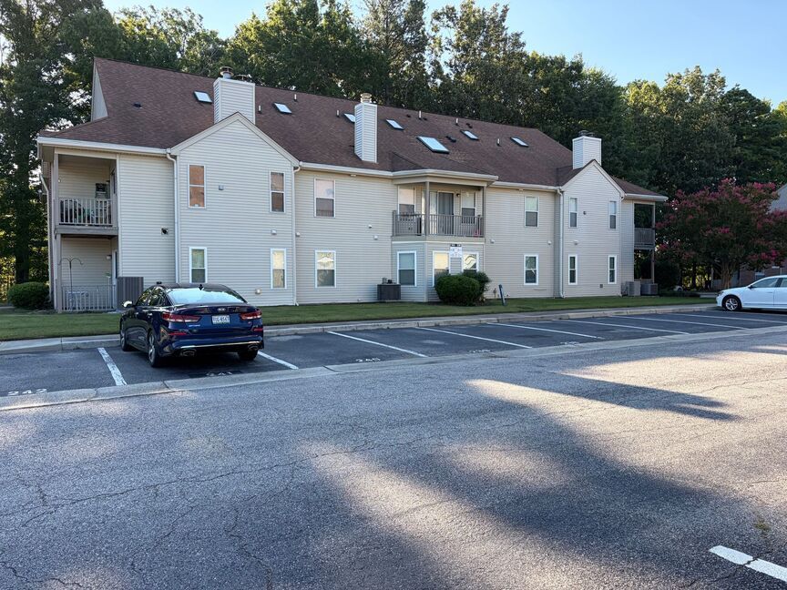 Apartment building with white siding, parked cars, and trees along a roadside.
