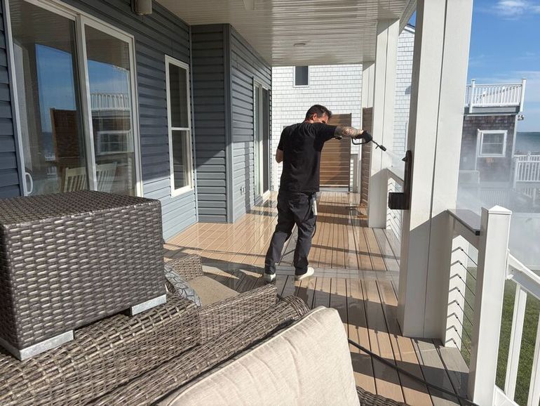 Person cleaning a sunlit wooden porch with white railings and wicker furniture