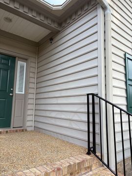 Front entrance with green door, beige siding, and black metal railing beside a gravel-covered porch.