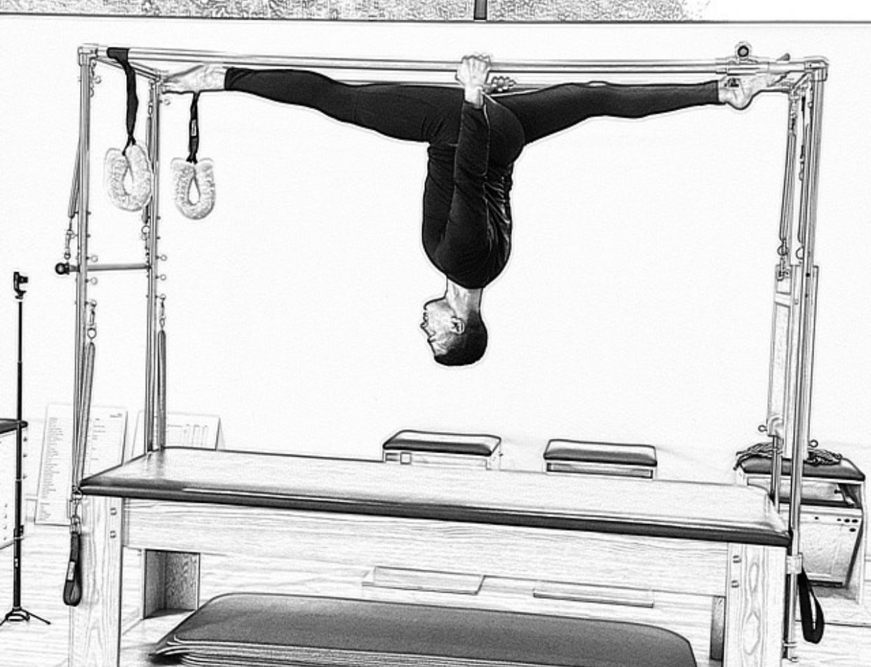 Person hanging upside down from a metal frame above a wooden table in a black-and-white studio photo