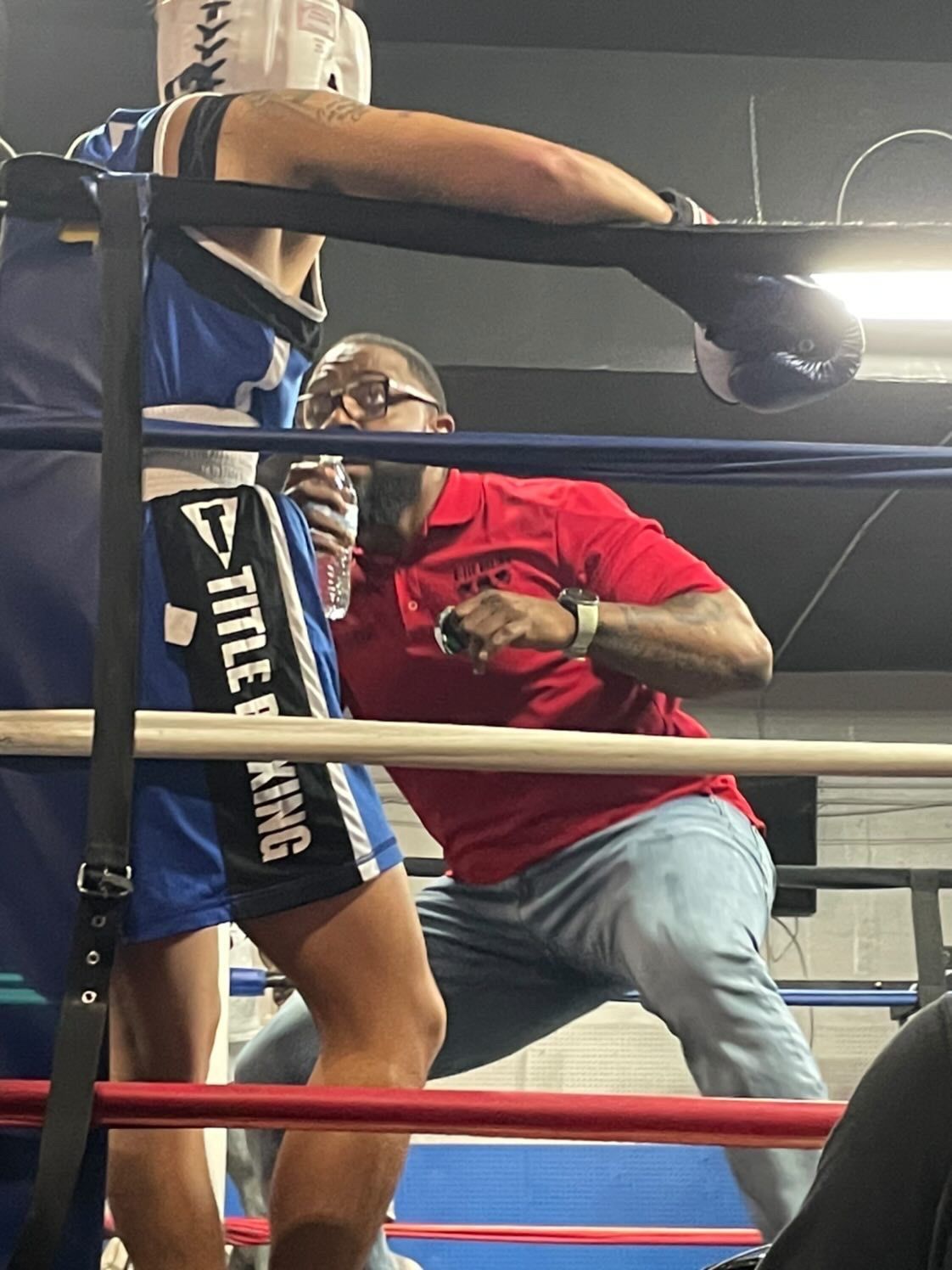 A man in a red shirt is watching two boxers in a boxing ring