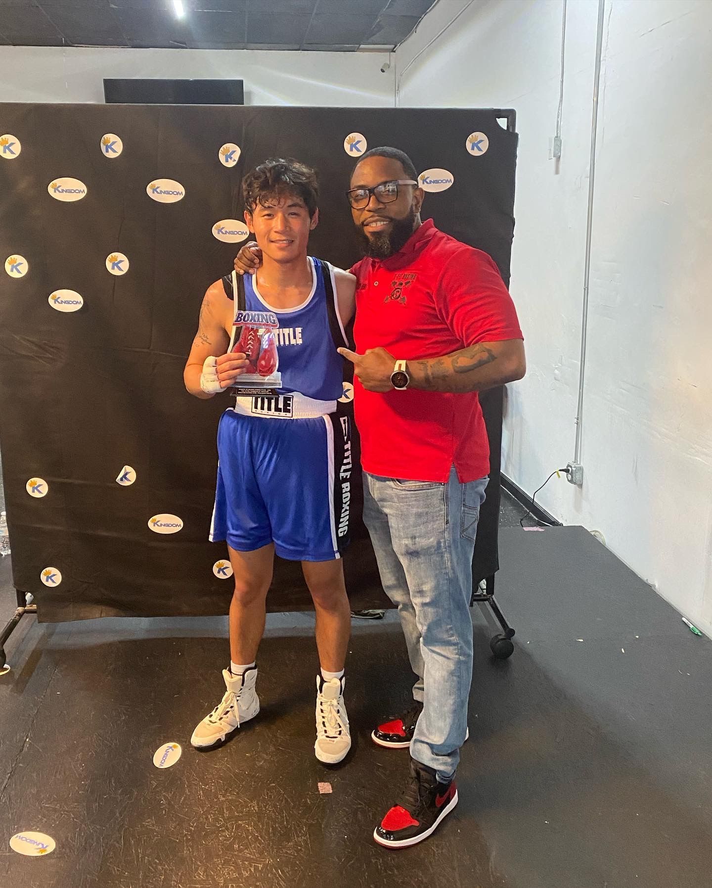 A man in a red shirt is standing next to a young man in a blue boxing uniform.