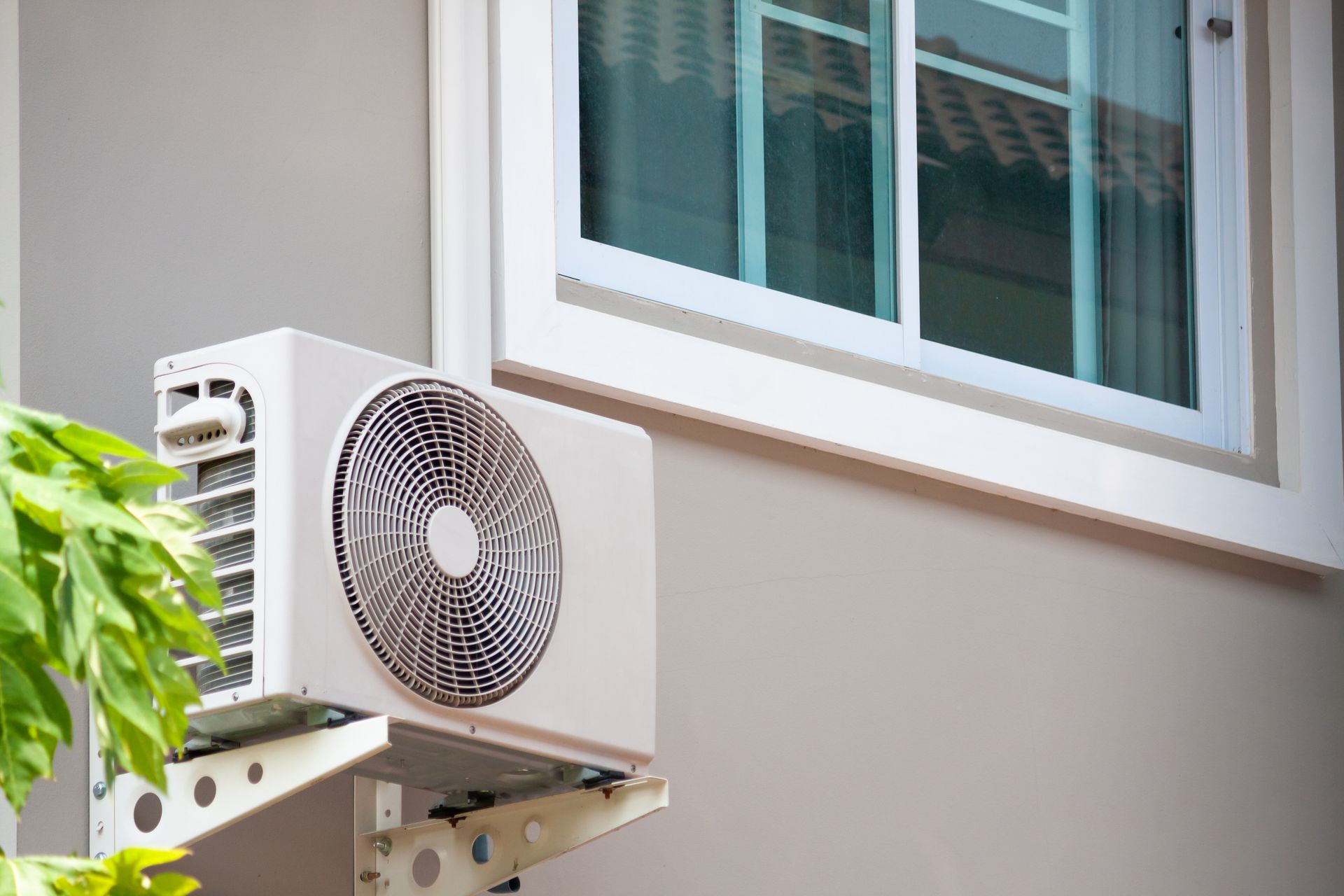 A white outdoor air conditioning unit mounted on a bracket on a beige house wall next to a window.
