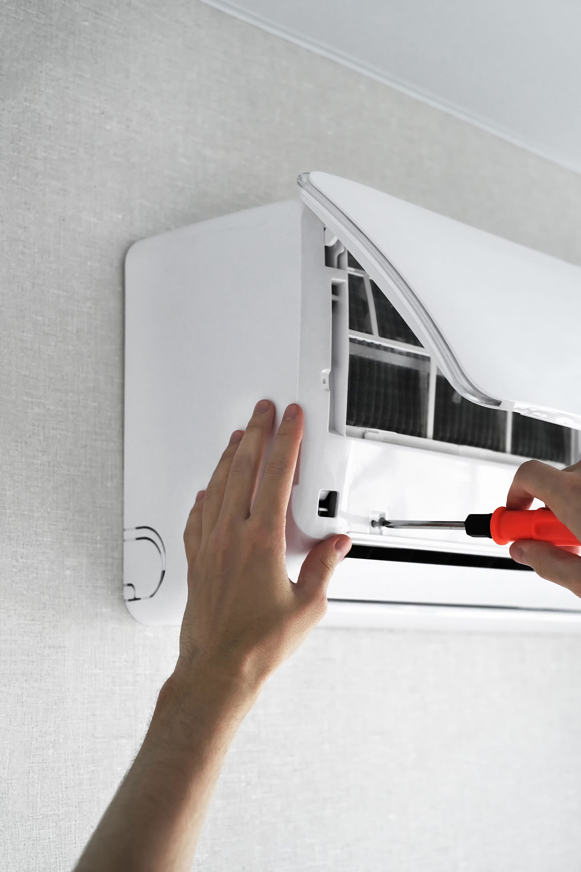 Hands using a red-handled screwdriver to repair or maintain a wall-mounted white air conditioning unit.