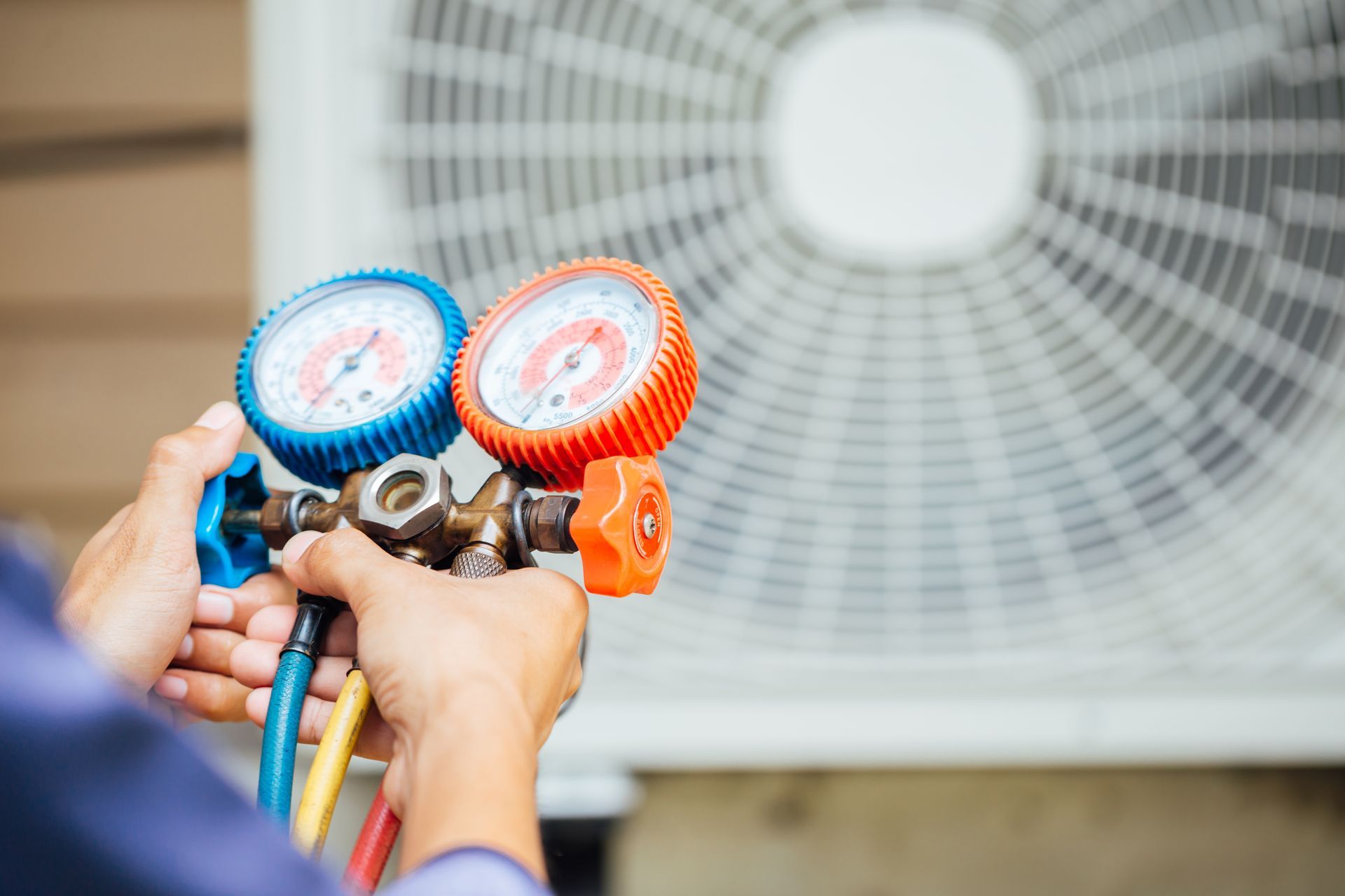 A technician holding a set of HVAC pressure gauges in front of an outdoor air conditioning unit.