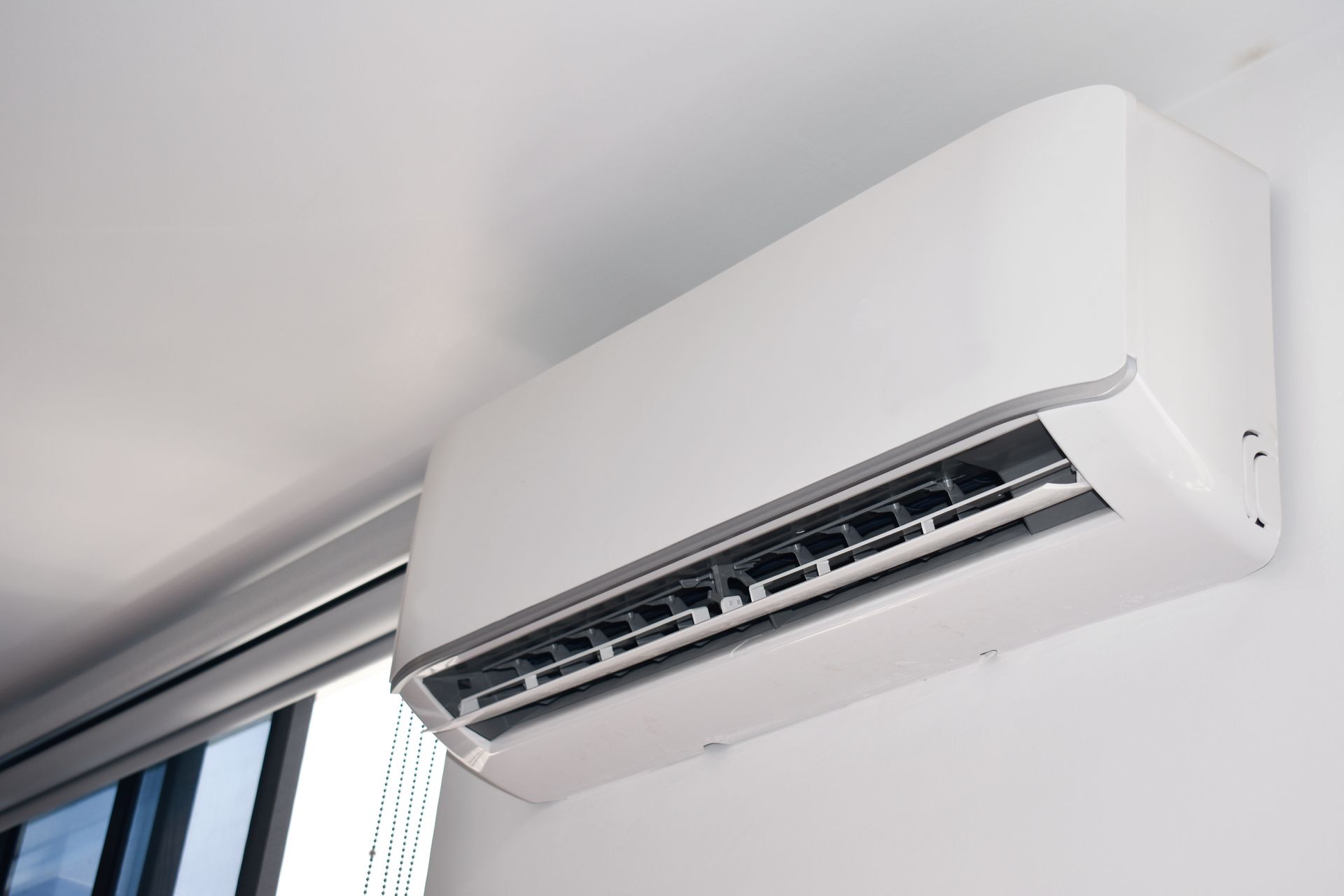 A white wall-mounted air conditioning unit installed high on a wall next to a window.