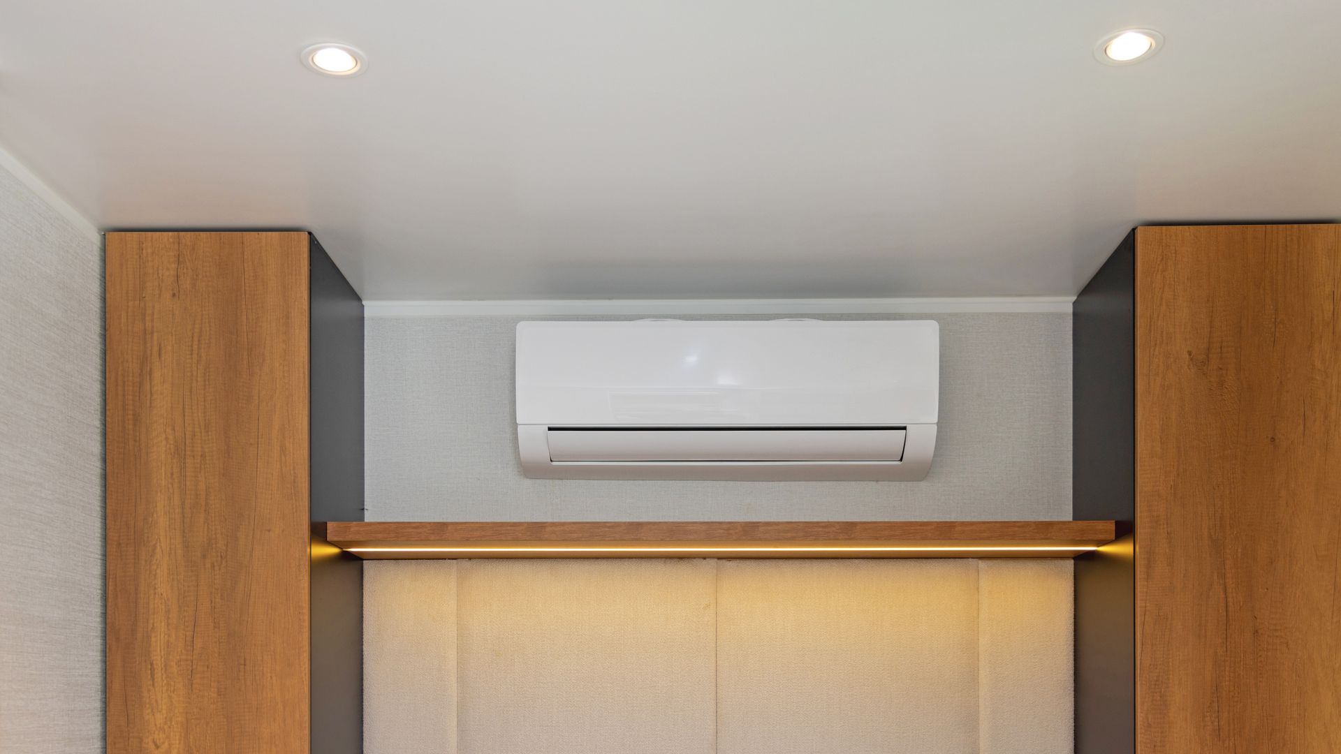 A white air conditioning unit mounted on a grey wall between two wooden cabinet units, with a shelf and warm lighting below.