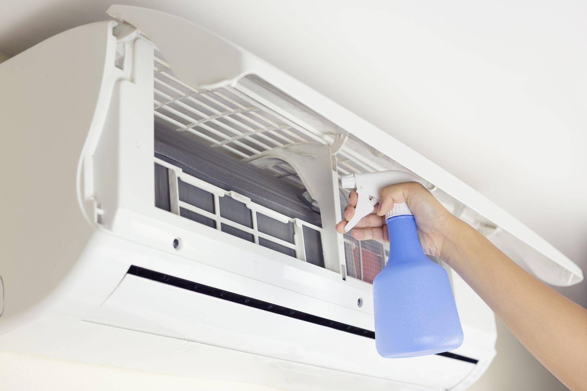 A hand sprays cleaning solution from a blue bottle onto the internal filter of a wall-mounted air conditioner.