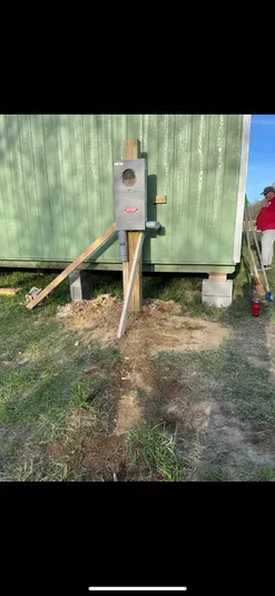 Electrical meter box on a wooden pole, supported by braces, in front of a green building.