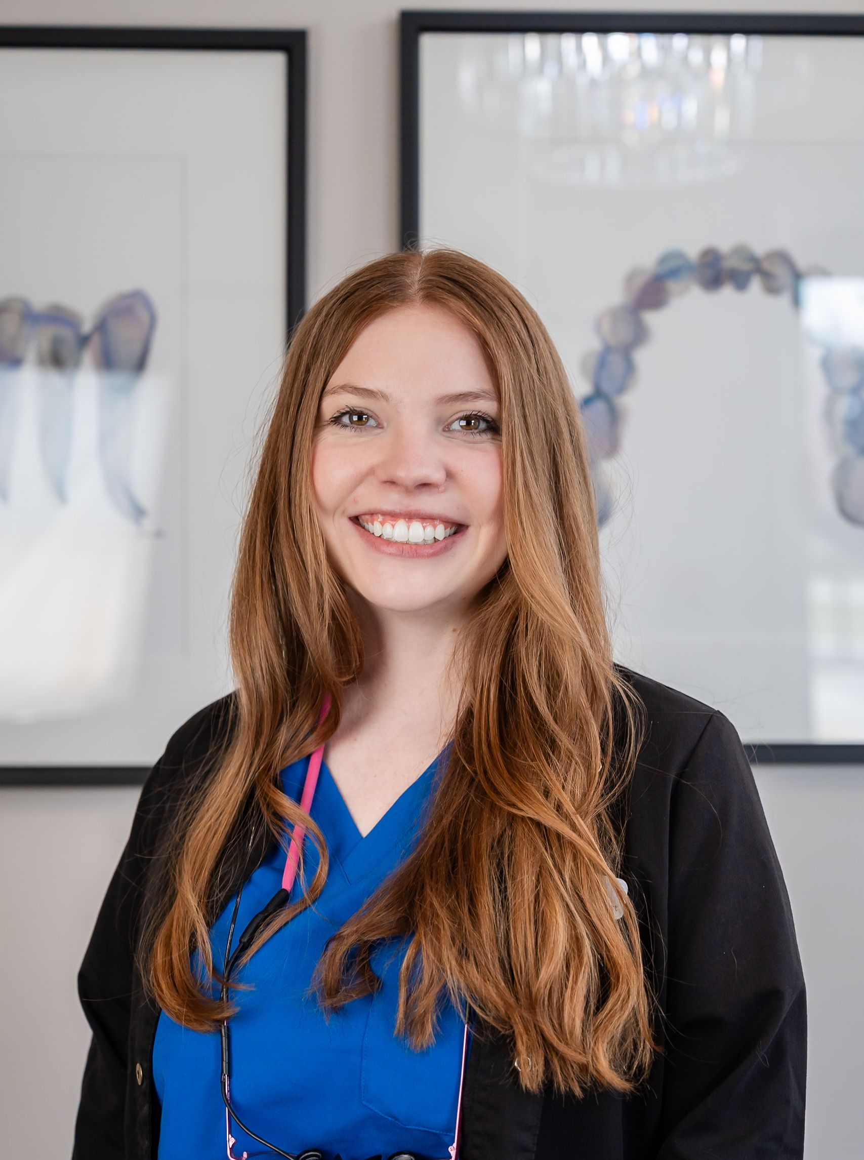 Smiling person in a blue blouse and black cardigan standing in an office with framed dental X-rays.