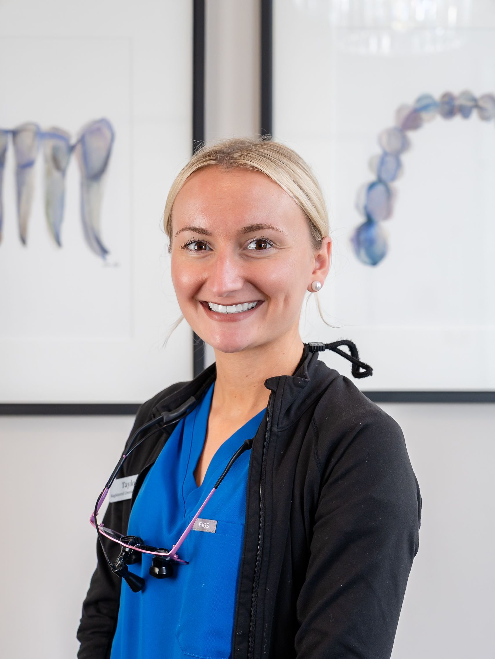 Smiling dental professional in blue scrubs with black jacket, standing in a clinic office.