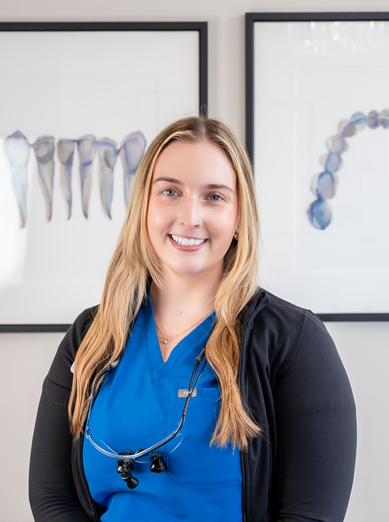 Smiling person in blue scrubs with black jacket, standing in a dental office with tooth anatomy posters.