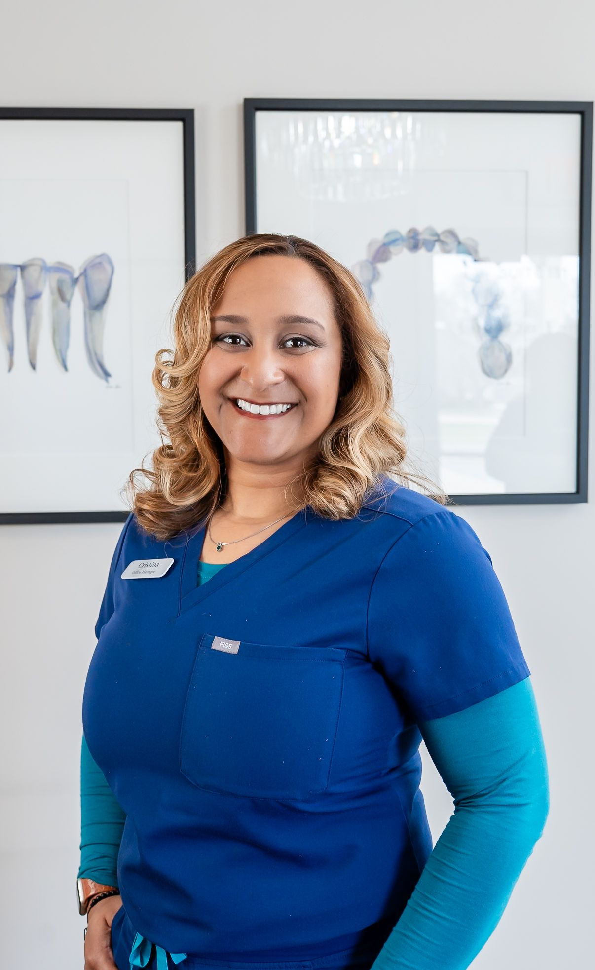Smiling staff member in blue uniform standing in an office with framed dental x-rays behind her