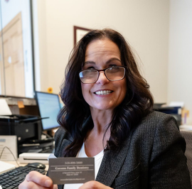 Smiling woman at a desk holding a business card in an office with a computer