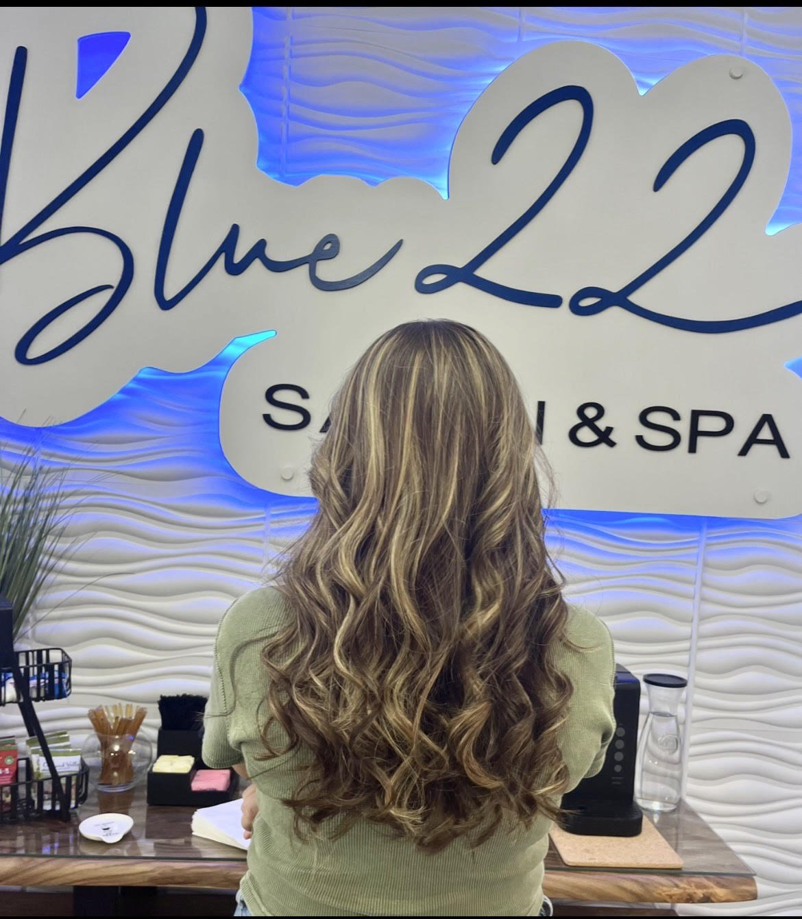 Woman with long wavy hair standing at a salon counter beneath a Blue 22 Spa sign