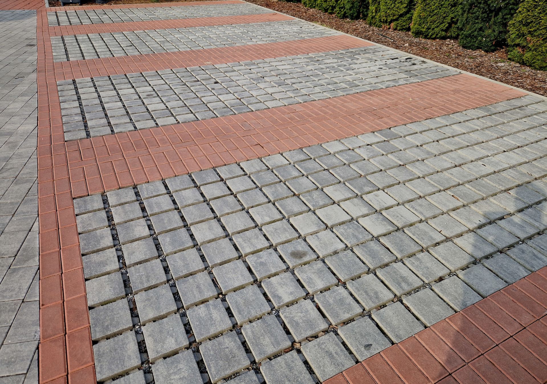 Patterned brick walkway with red borders and gray square pavers beside shrubs