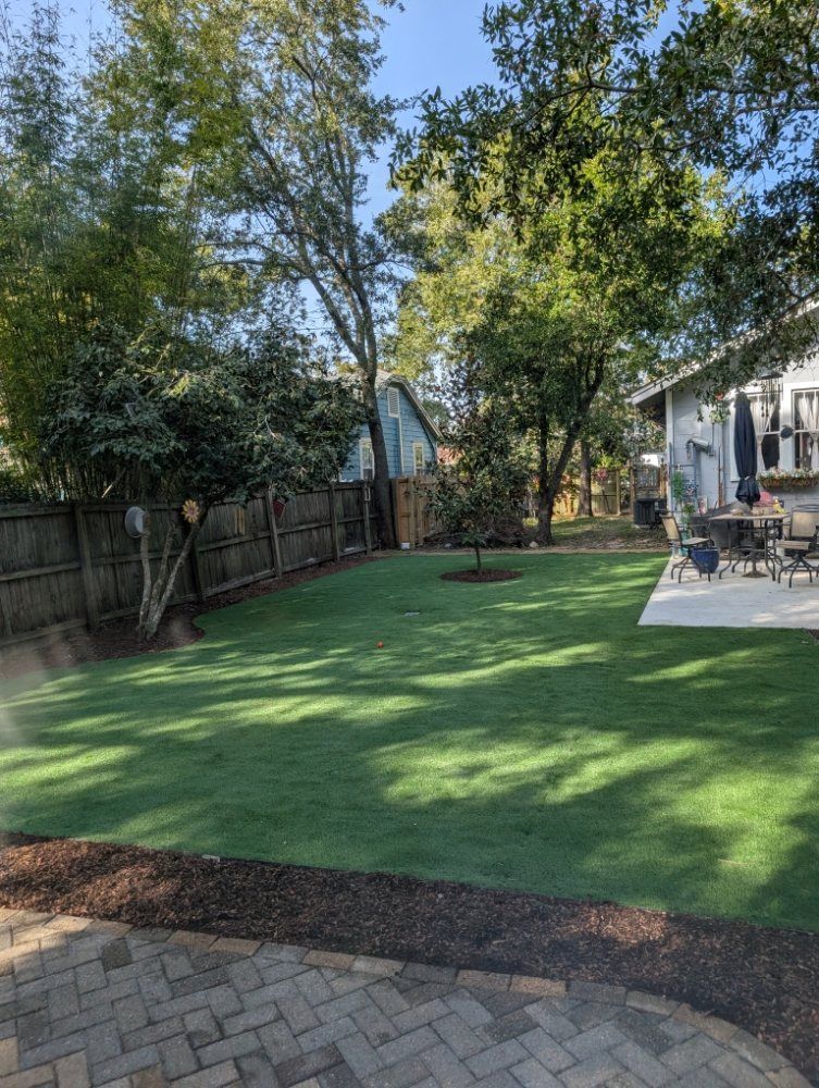 Backyard with artificial turf, trees, wooden fence, and a patio beside a house
