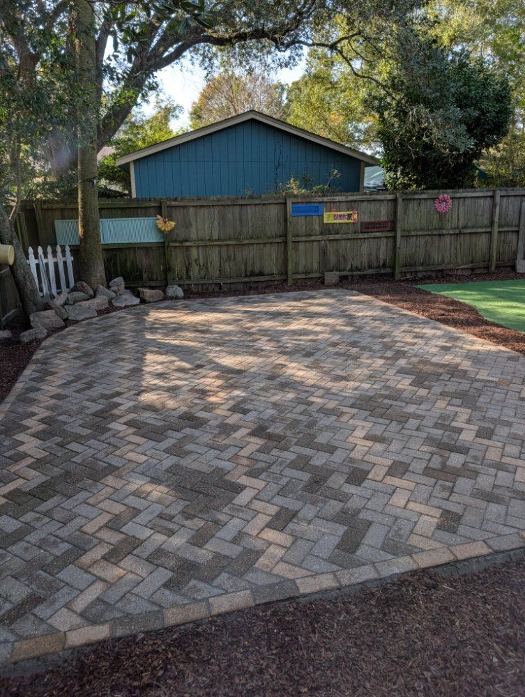 Brick-paved driveway in front of a fenced backyard with trees and a blue shed