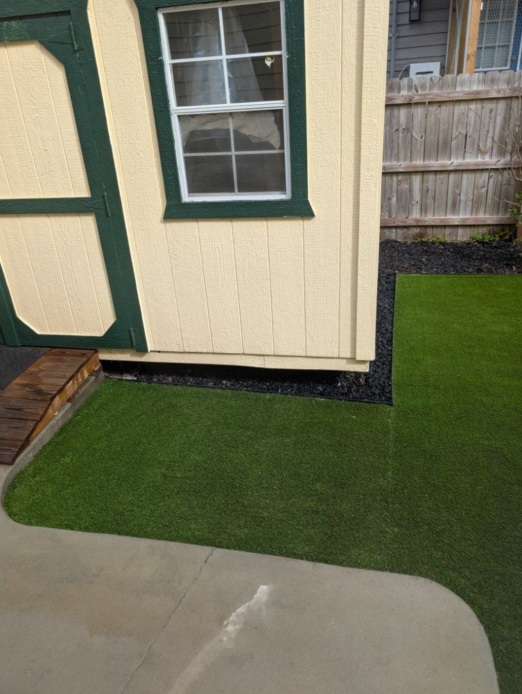 House corner with window, beige siding, green trim, and artificial turf beside a concrete patio.