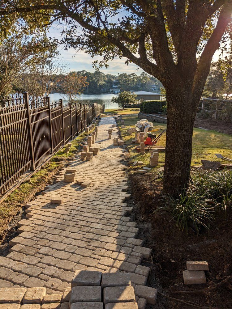 Brick path beside fence and tree leads to a lake, with a person crouching near the water.
