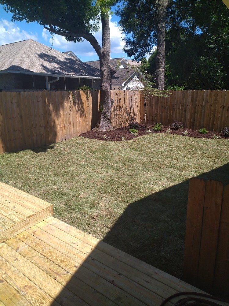 Backyard with wooden deck, grass lawn, tall fence, and trees in sunlight