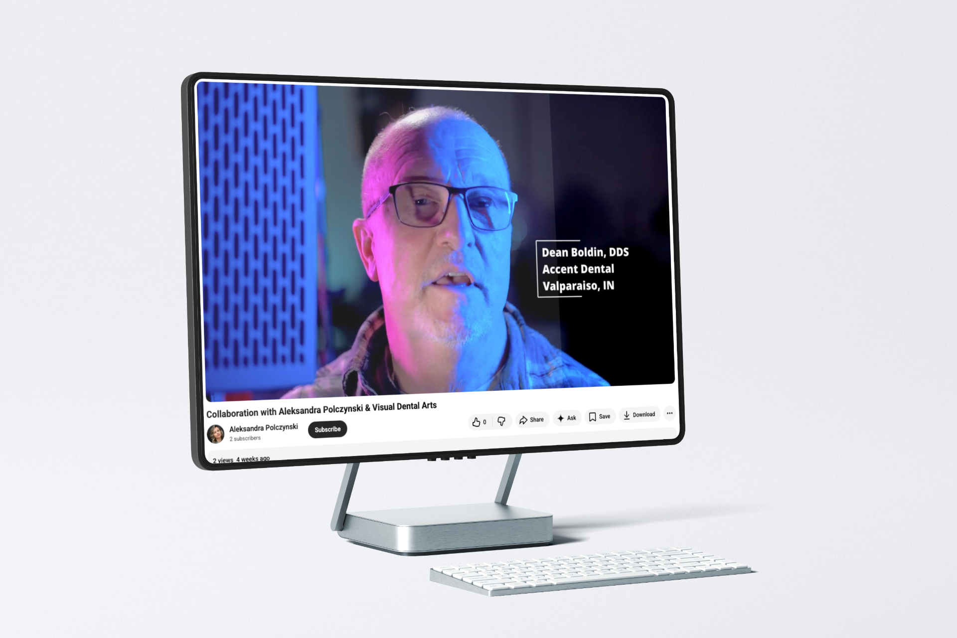 Computer monitor displaying a video of a man speaking, lit with blue and pink lights. White keyboard below.