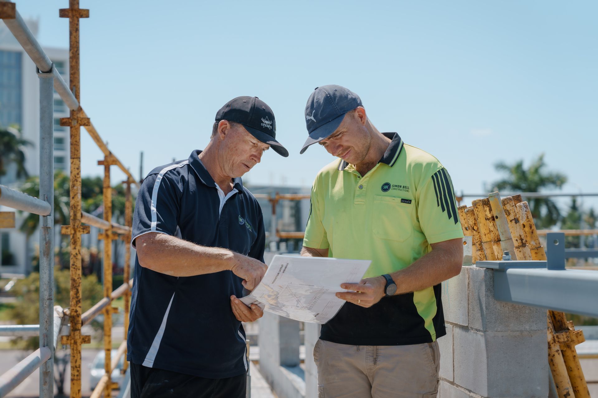 Two Men In Caps, On A Rooftop, Reviewing A Blueprint. One Points To The Document. Sunny Day — Simon Bell Constructions in Alice River, QLD