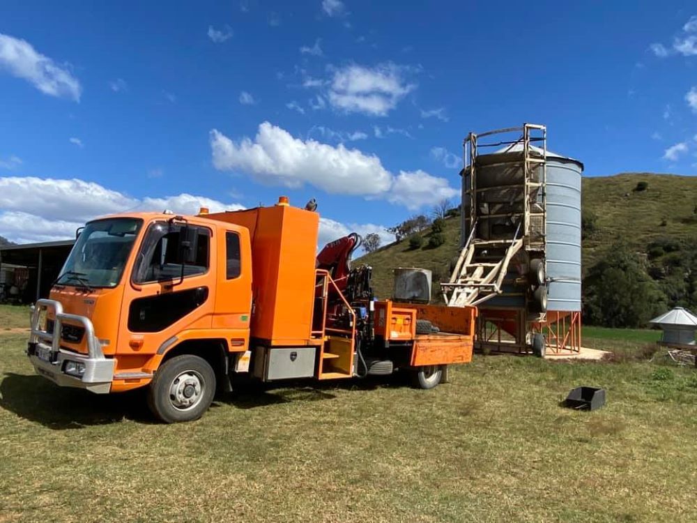 An Orange Truck is Parked in a Grassy Field Next to a Silo — Wallamore Grain & Produce In Wallamore, NSW