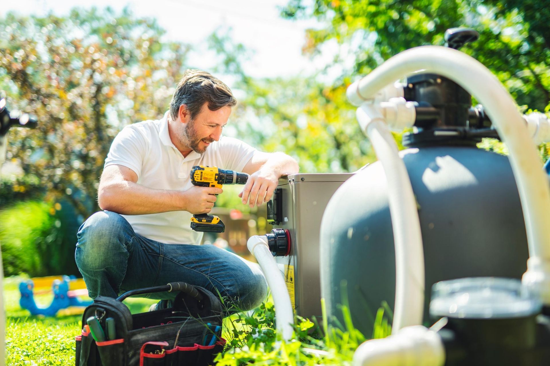 Man using a drill to work on outdoor plumbing beside large pipes in a sunny yard