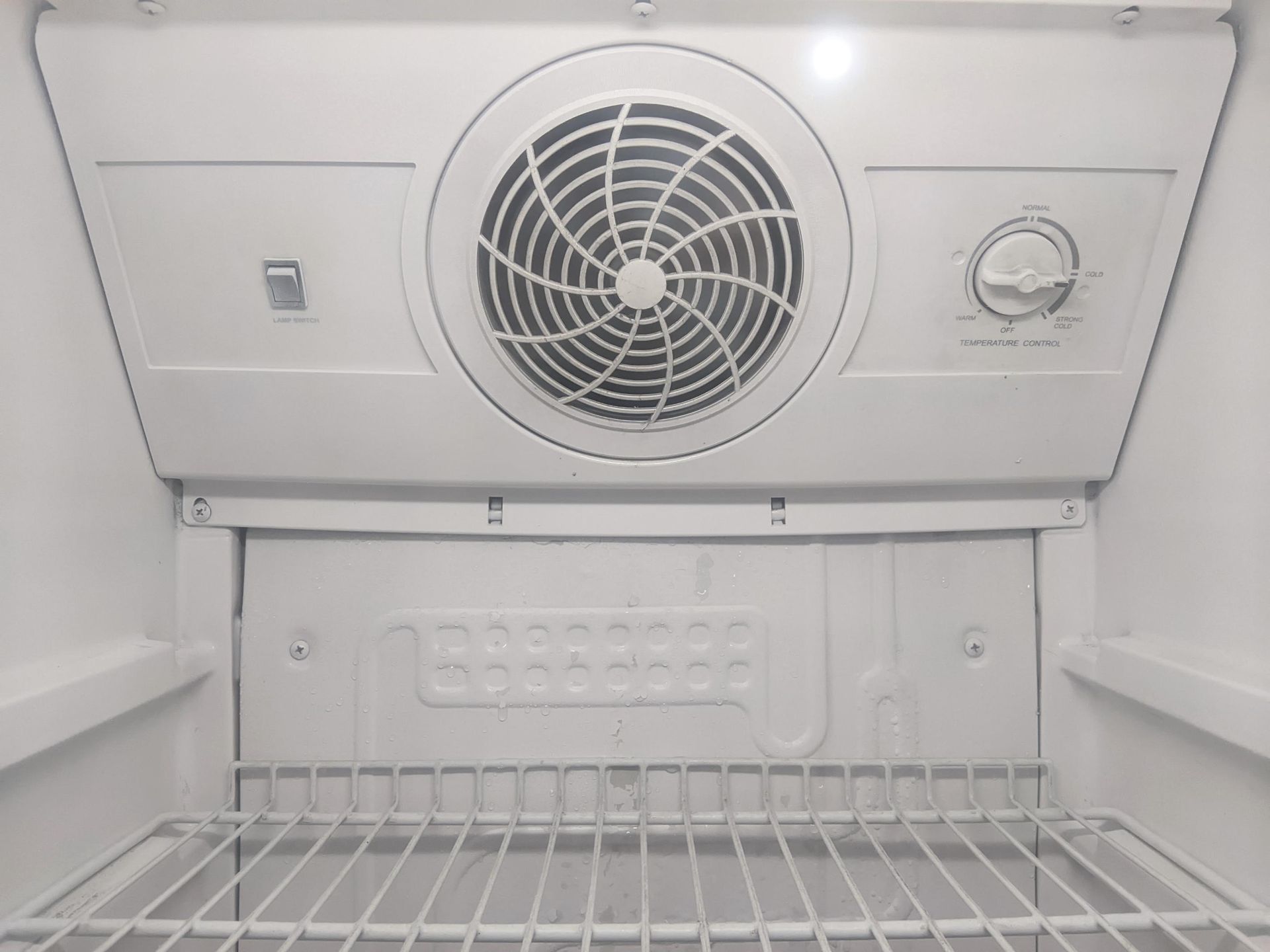 Empty white refrigerator interior with wire shelf and circular fan vent at the back.