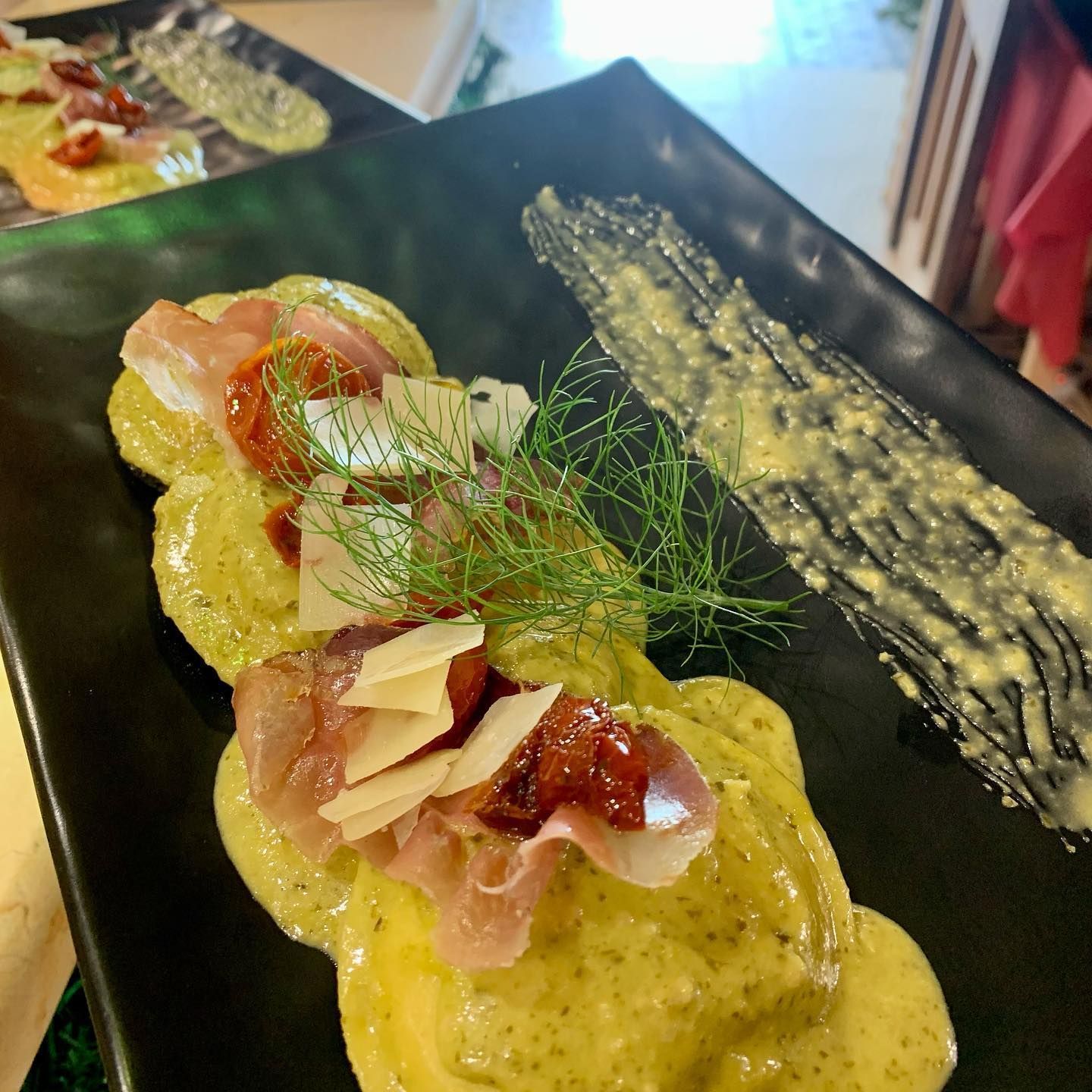 Ravioli serviti su un piatto rettangolare nero, conditi con prosciutto crudo, pomodori secchi, parmigiano e finocchio, con salsa alle erbe aromatiche.