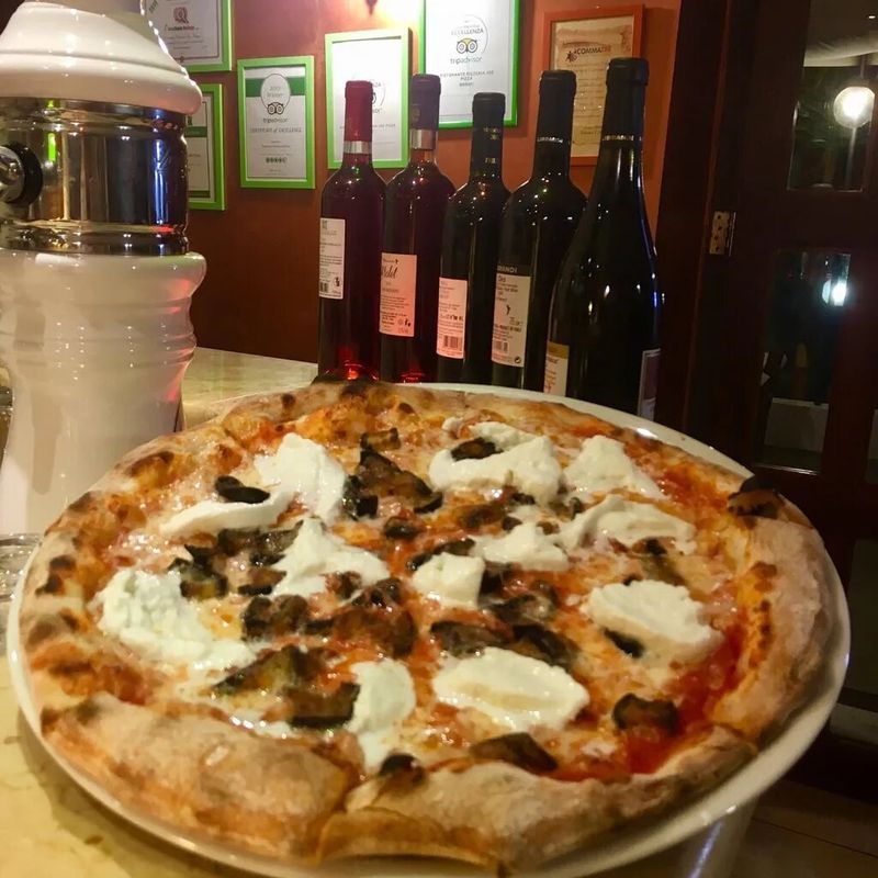 Una pizza con formaggio fuso e melanzane servita su un piatto bianco in un ristorante con bottiglie di vino sullo sfondo.