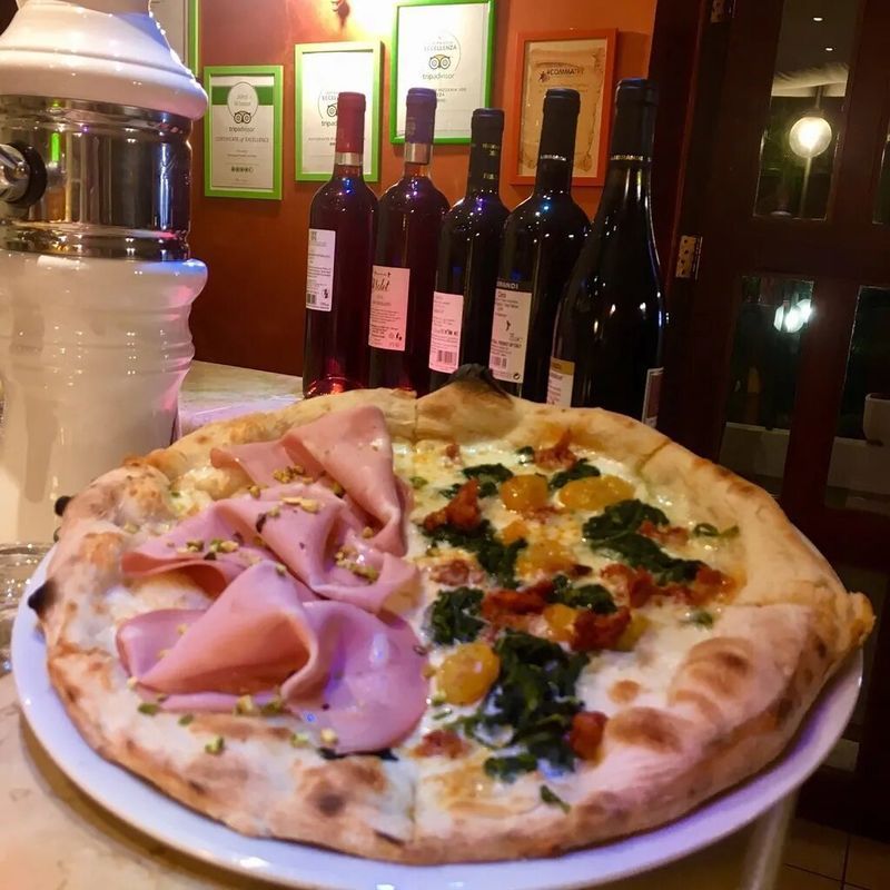 Una pizza metà mortadella e metà verdure servita su un piatto davanti a una fila di bottiglie di vino in un ristorante.