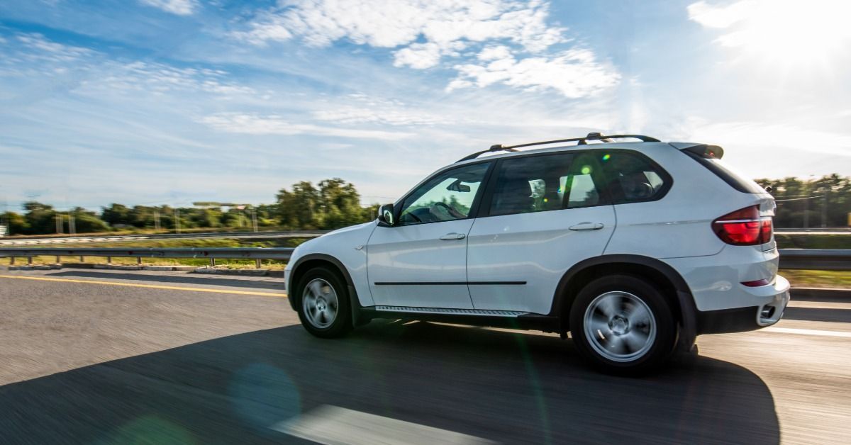 A white car driving alone on the highway during the daytime. Many tall trees are visible in the distance.