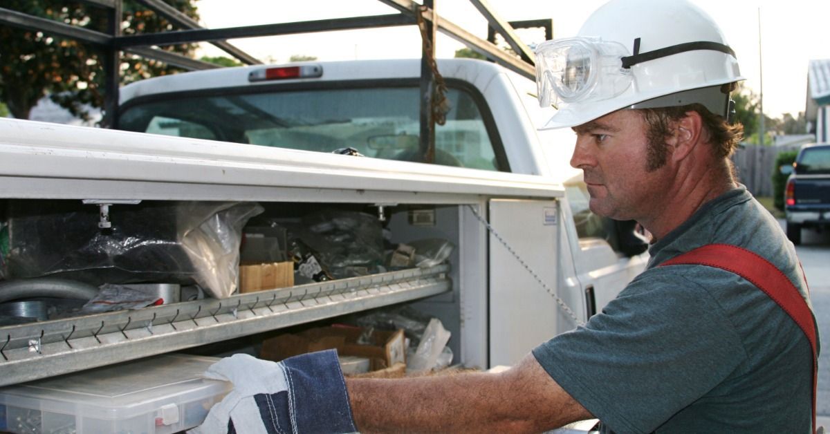 An electrician wearing a white hard hat and work gloves removing a small plastic box from their work truck.